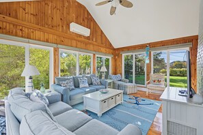 A spacious living room with a high ceiling, wooden paneling, a ceiling fan, and large windows offering a view of the outdoors. - New to our 2027 Inventory!   Spacious 2700 sq. ft.  home with heated pool! (West Yarmouth)