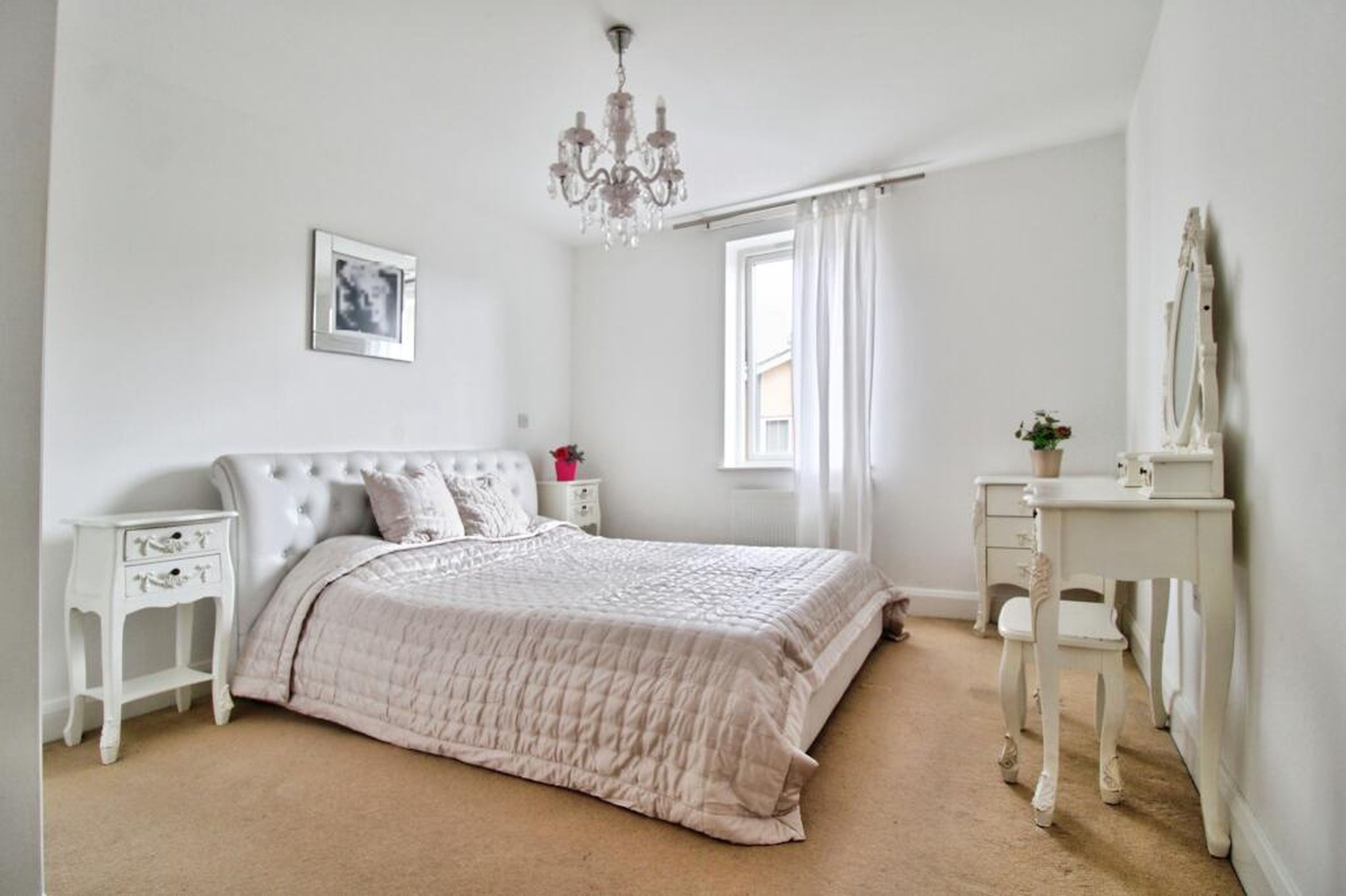 A bedroom with a bed, bedside tables, a dresser, a chandelier, a window with curtains, and a picture on the wall.