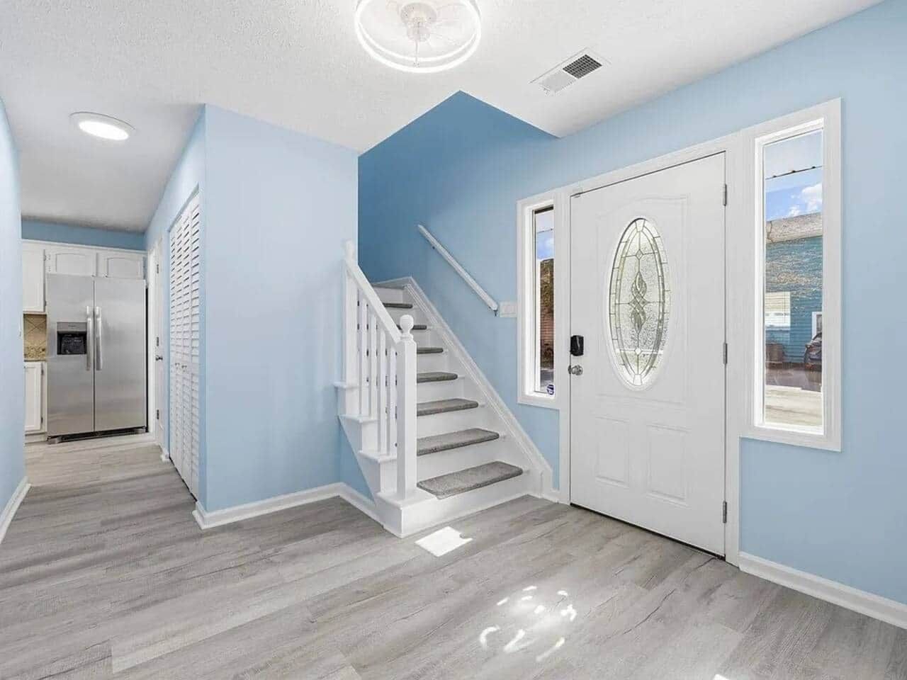 A hallway with a staircase, a white door with a glass window, and a kitchen area in the background.