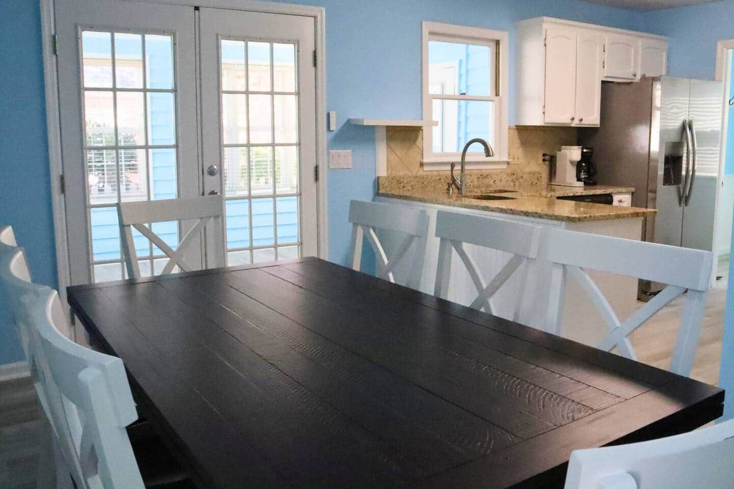 A dining area with a dark wooden table and white chairs, a kitchen with white cabinets and stainless steel appliances, and a window with a view of the outside.