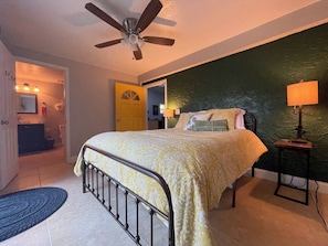 Une chambre à coucher comprenant un lit, une table de chevet, un ventilateur de plafond et une porte donnant sur une autre pièce.