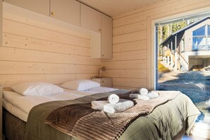 A wooden cabin room with a bed, pillows, a bedside lamp, and a view of a wooden structure outside. - Alpine South Slope A7 by Aavalevi (Levi)