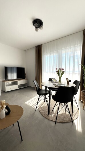 Ein modernes Wohnzimmer mit einem Flachbildfernseher, einem Esstisch mit Stühlen und einem großen Fenster mit Vorhängen.