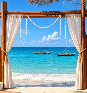 Una estructura de madera con cortinas blancas y cuentas colgantes, con vista a una playa con barcos y agua azul cristalina.