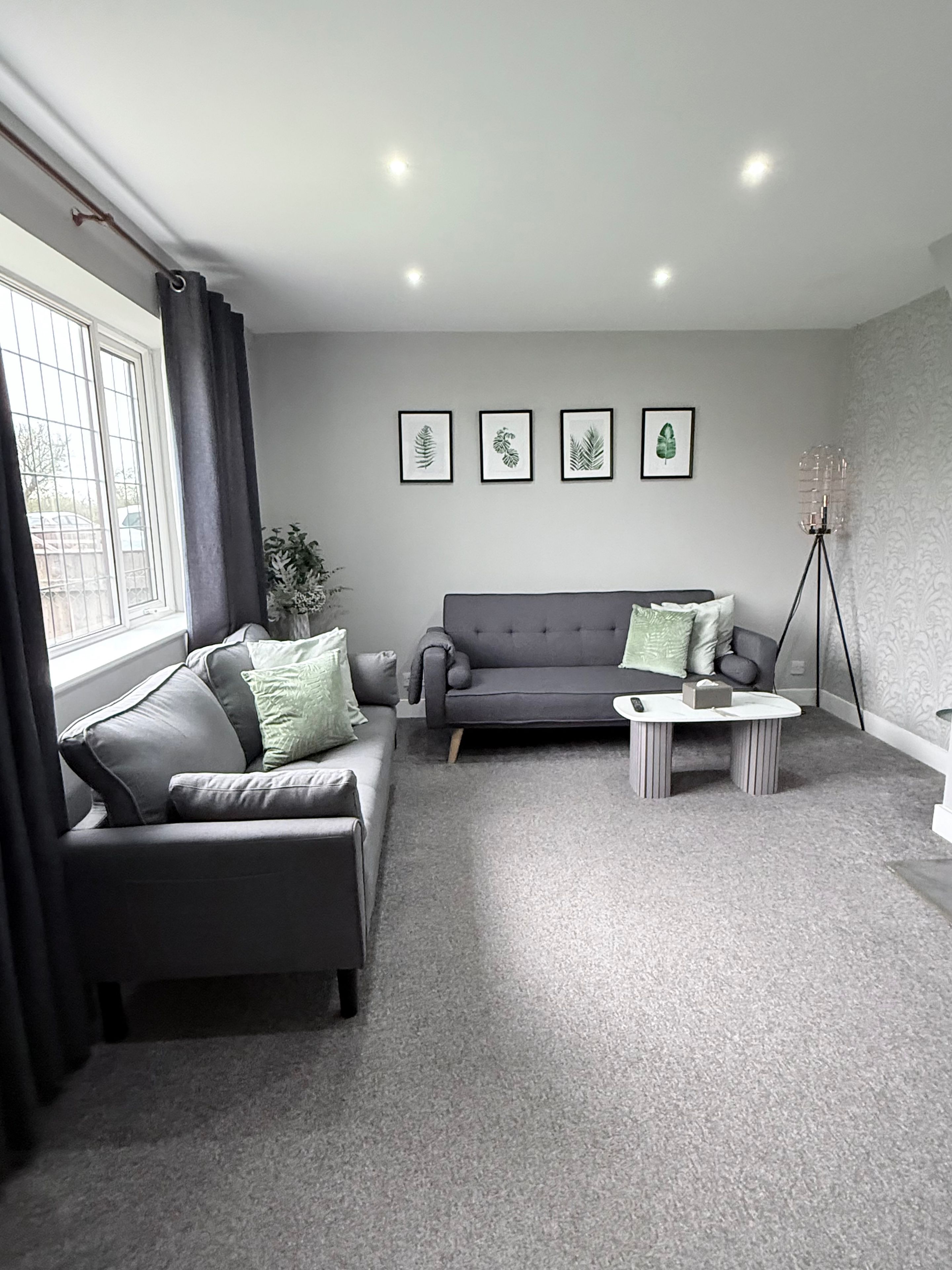 Ein modernes Wohnzimmer mit einem grauen Sofa, einem Couchtisch und einer Stehlampe. Vier gerahmte Blattdrucke sind an der Wand angebracht.