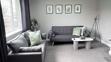 Una sala de estar moderna con un sofá gris, una mesa de centro y una lámpara de pie. Hay cuatro láminas enmarcadas con motivos de hojas en la pared.