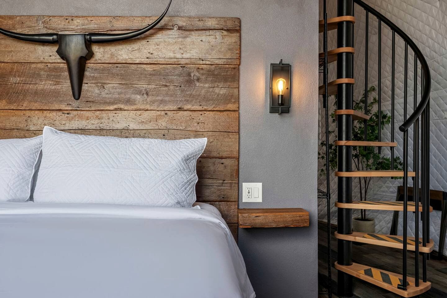 A bedroom with a wooden headboard, a bed with white bedding, a wall-mounted light, and a spiral staircase.