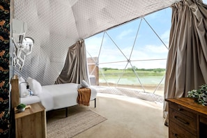 Ein Schlafzimmer mit Bett, Nachttisch, Lampe und Blick auf ein Gewässer durch ein großes Fenster.