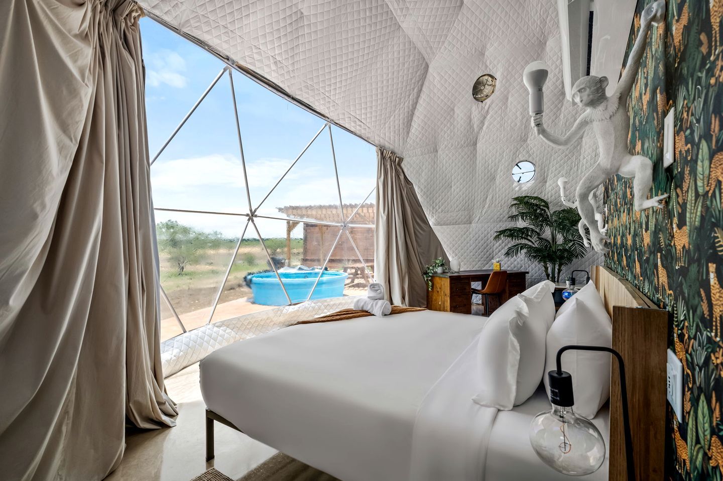 A room with a bed, a desk, a chair, a clock, a light fixture, and a view of an outdoor area with a pool.