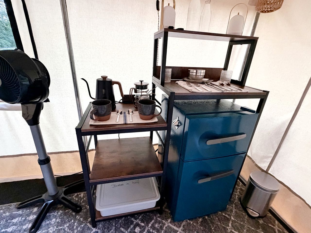 Una kitchenette con cafetera, hervidor y utensilios sobre una estantería, un armario azul con cajones y un cubo de basura.
