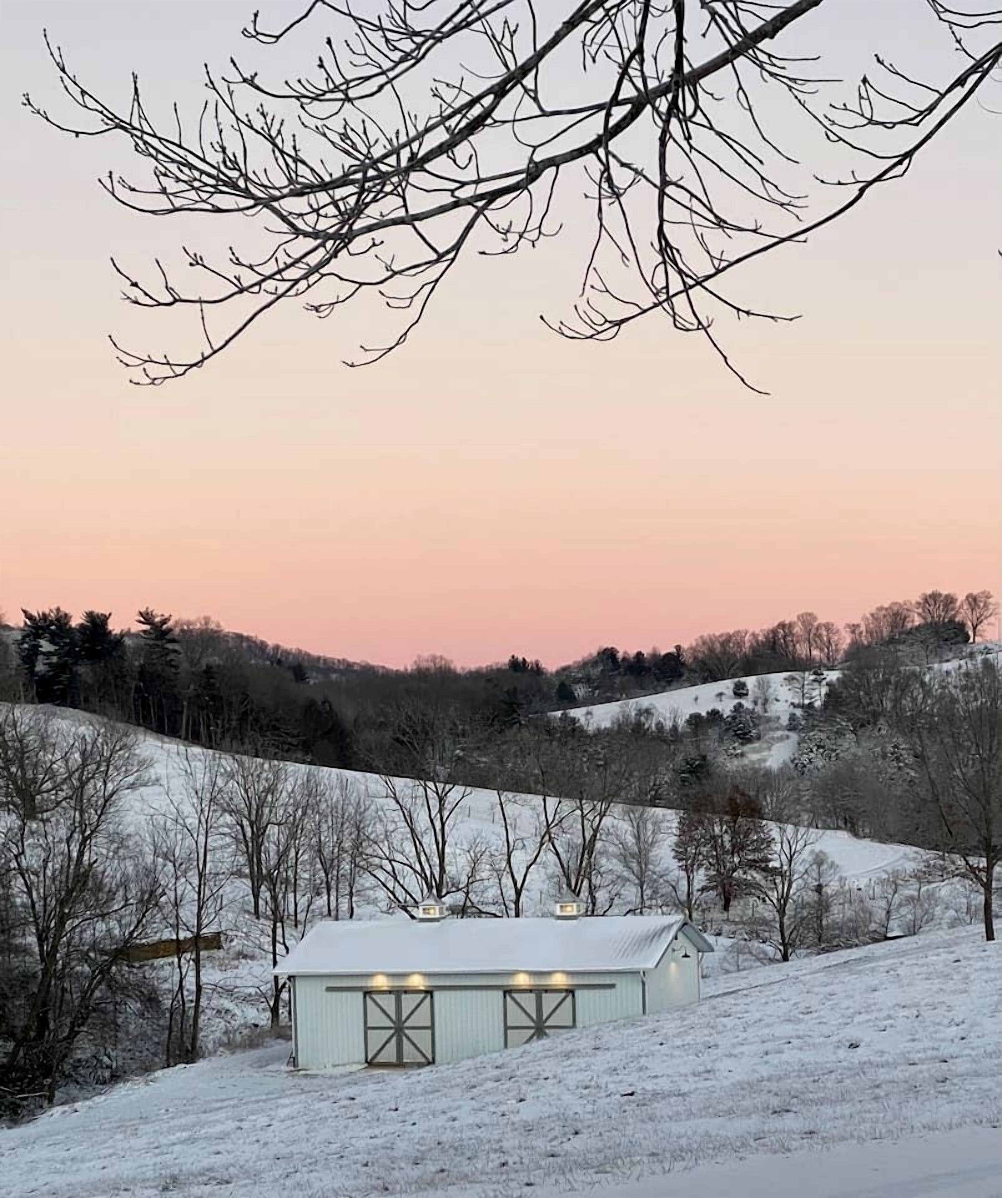 Schneebedeckte Landschaft mit einem kleinen Gebäude, das eine Scheunentür und Beleuchtung aufweist, umgeben von kahlen Bäumen und Hügeln.