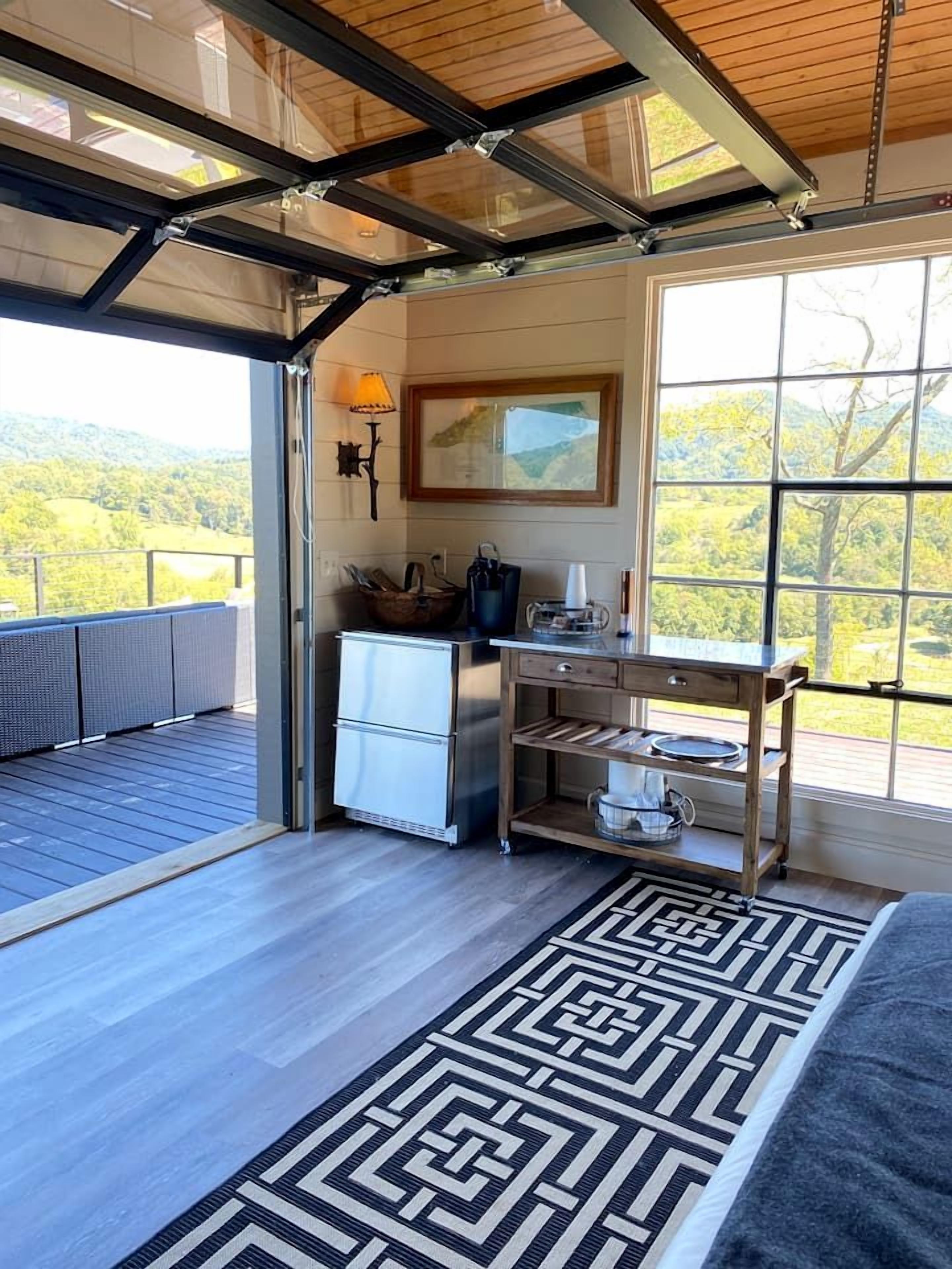 Una habitación con techo de madera, un ventanal con vistas a la vegetación, un refrigerador blanco y una mesa de madera con una lámpara.
