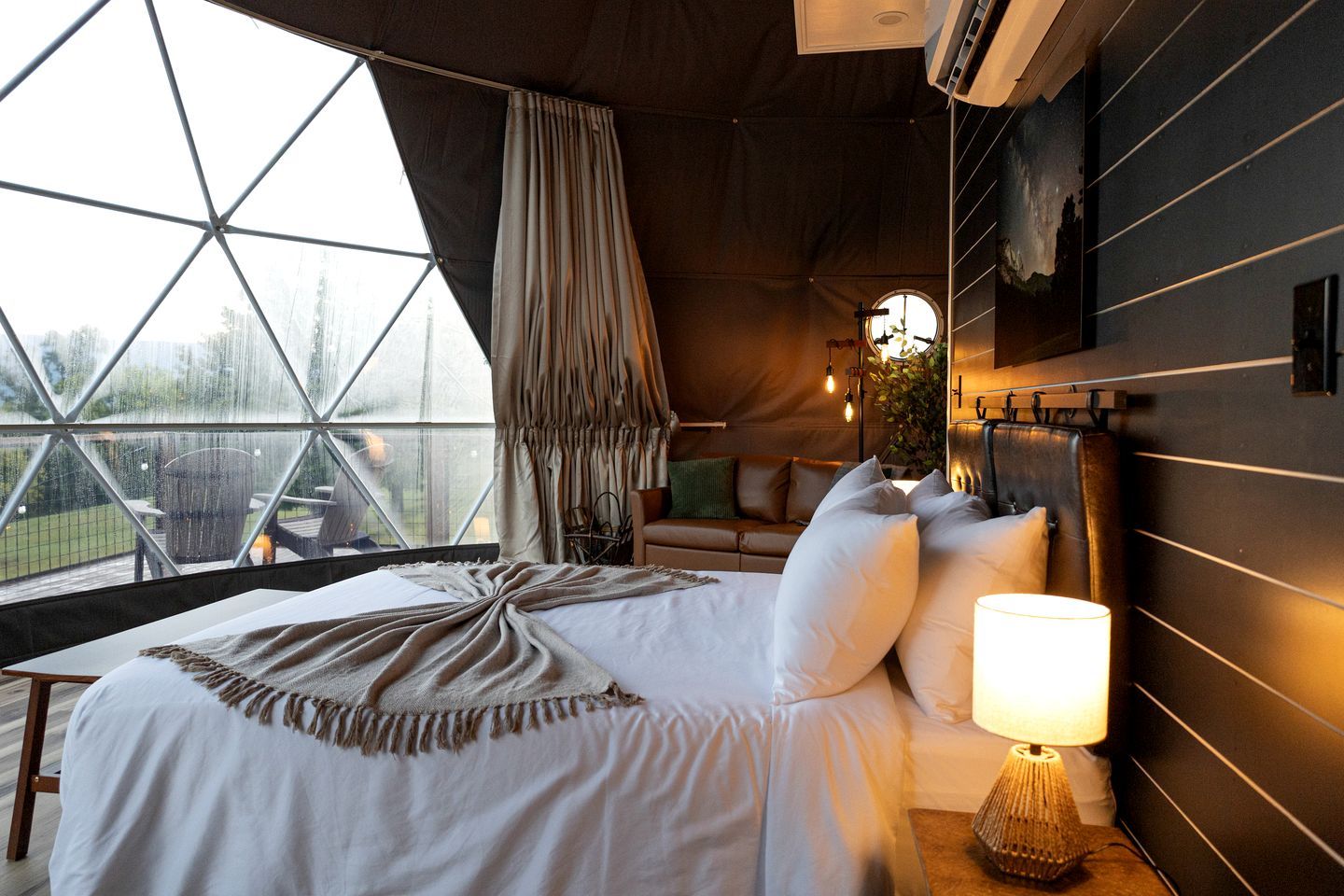 A bedroom with a large bed, a bedside lamp, a window with curtains, and a view of the outdoors.