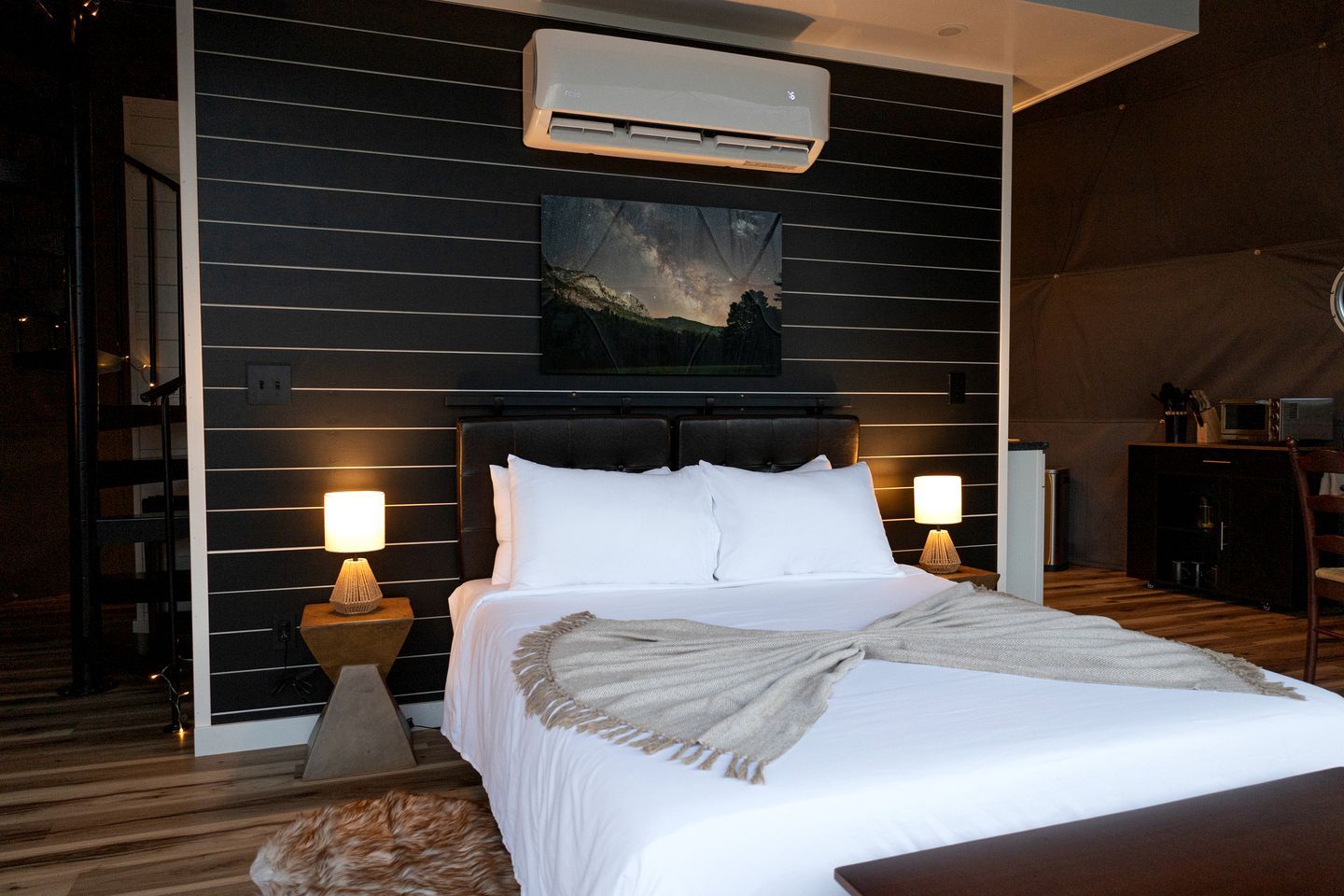 A bedroom with a bed, two bedside lamps, a wall-mounted air conditioner, and a painting above the bed.