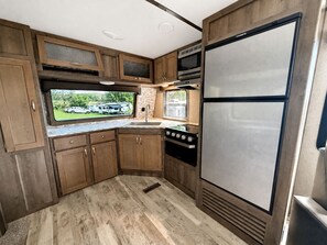 A compact RV kitchen with wooden cabinets, a stainless steel refrigerator, and a stove. - Spacious Rental Camper with Private Bathroom near Lake Champagne, Randolph, Vermont (Randolph)