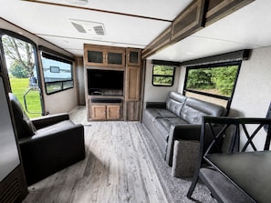 A compact RV interior with a seating area, a kitchenette, and a large window offering a view of greenery. - Spacious Rental Camper with Private Bathroom near Lake Champagne, Randolph, Vermont (Randolph)