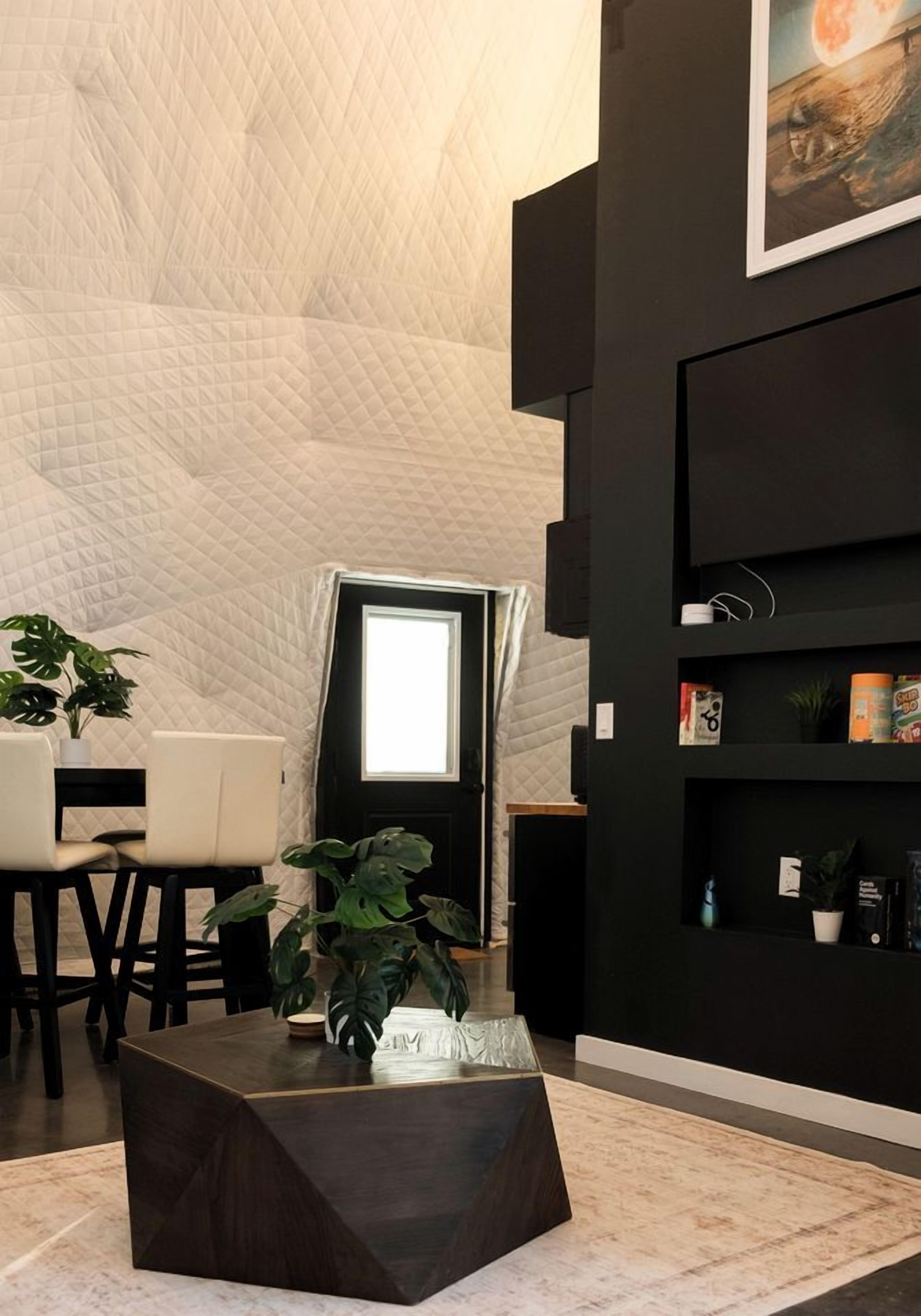 A modern living room with a geometric patterned wall, a black coffee table, a dining area with chairs, and a bookshelf with various items.