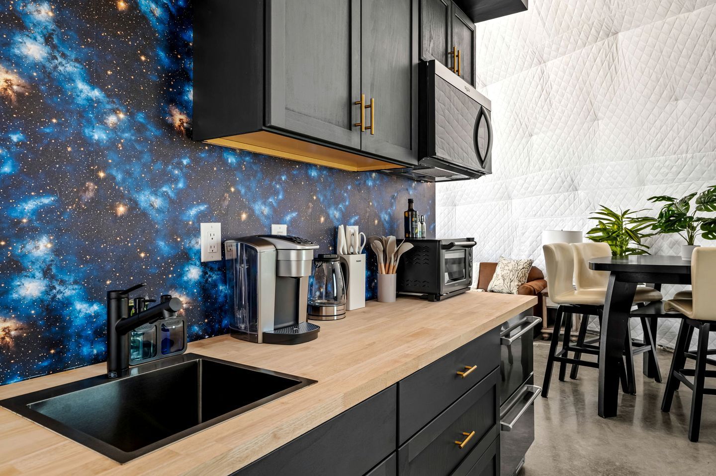 A kitchen with a galaxy-themed wall, a coffee machine, a microwave, and a dining area with chairs and a table.