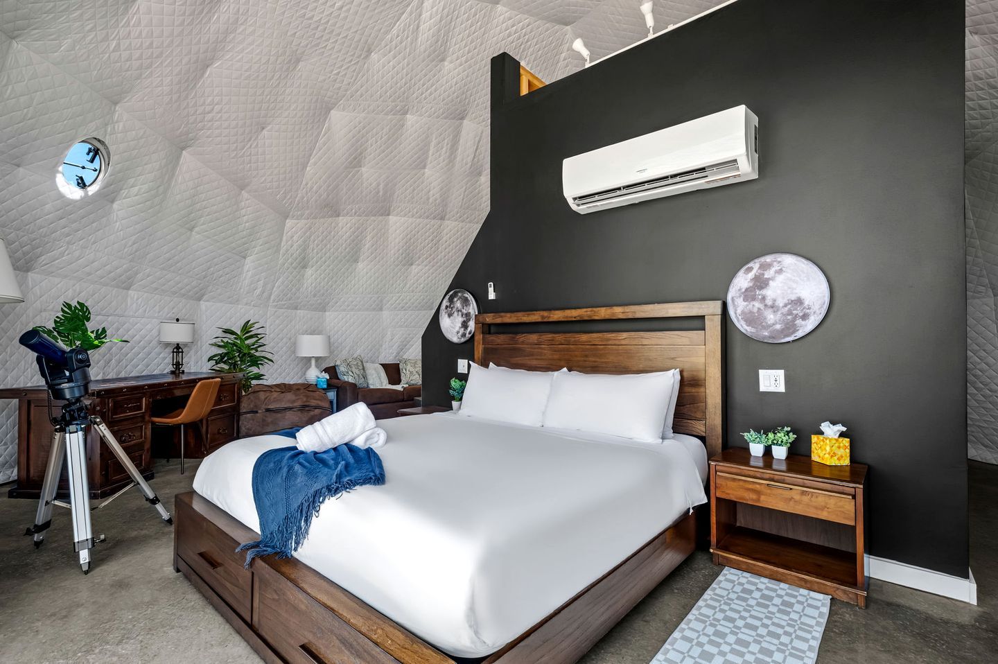 A bedroom with a large bed, a desk with a telescope, a wall-mounted air conditioner, and a round moon decoration.