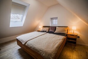 Ein Schlafzimmer mit schrägen Wänden, einem Bett mit Holzgestell, zwei Nachttischen mit Lampen und einem kleinen Fenster über dem Bett.