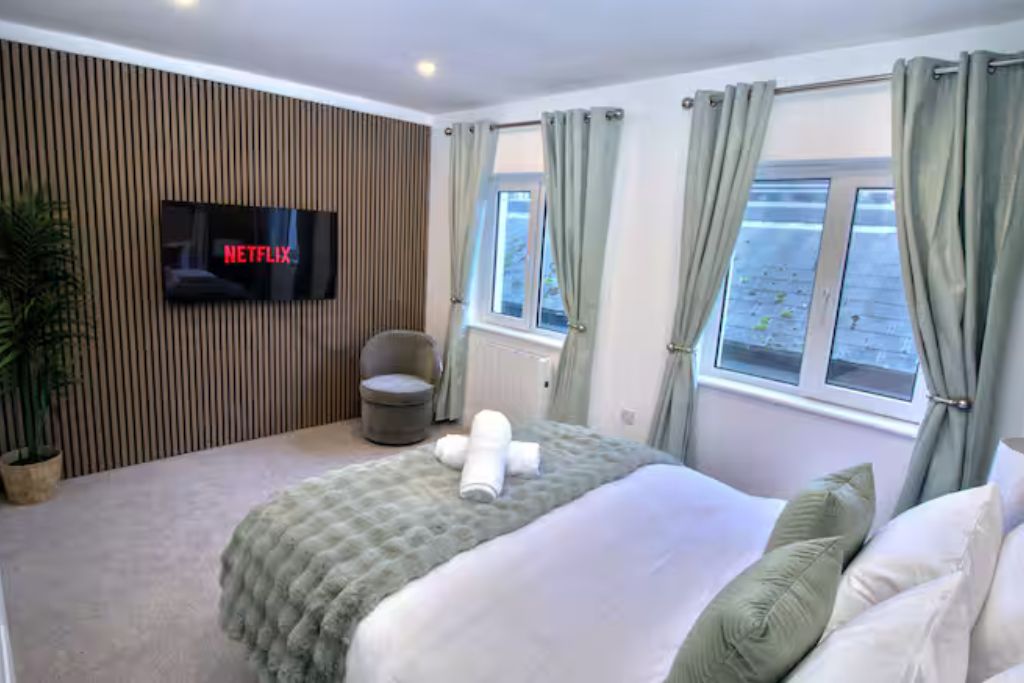 Una habitación de hotel con una cama, un televisor montado en la pared, una ventana con cortinas y una planta en maceta.