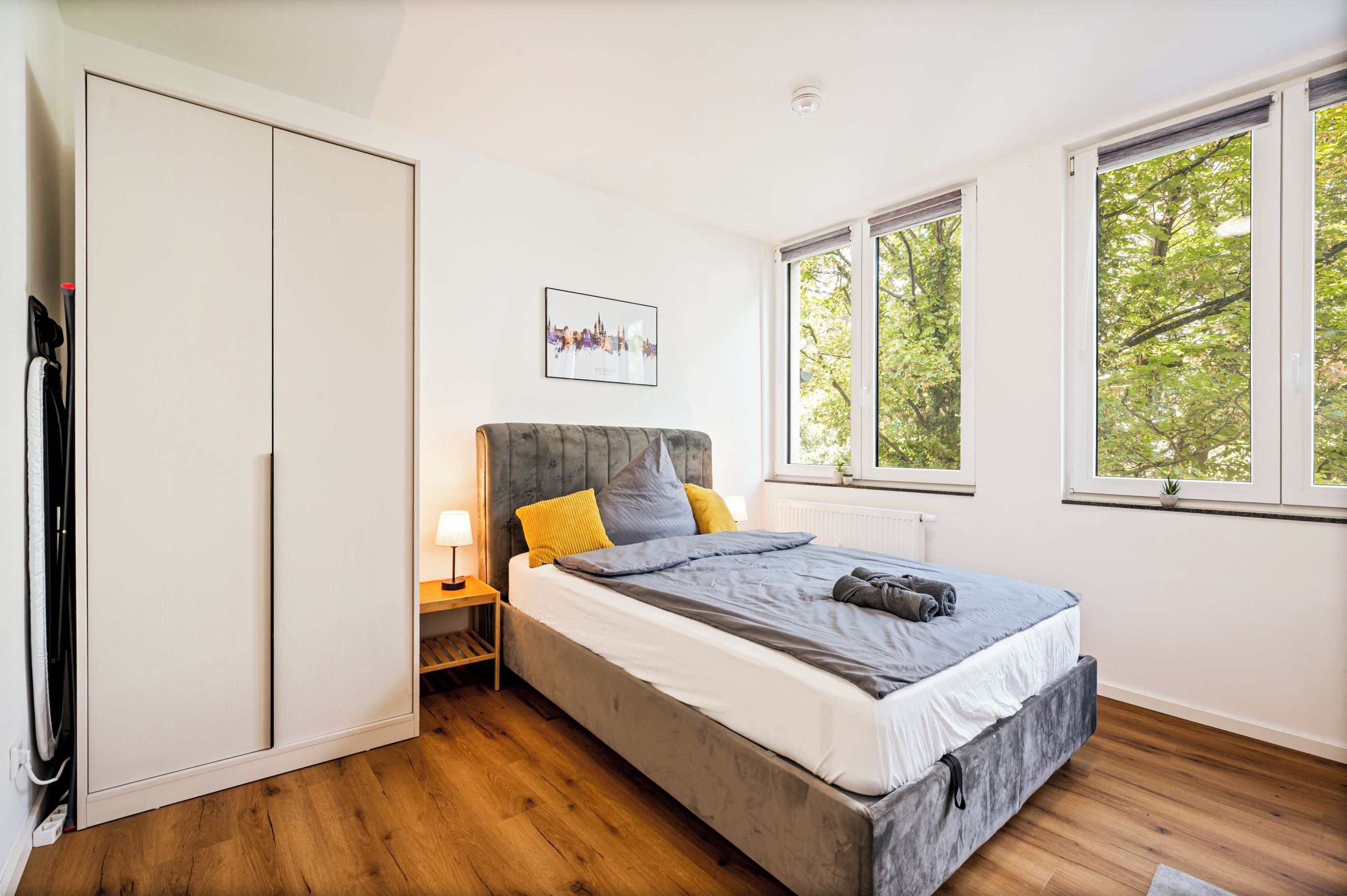 A bedroom with a bed, bedside table, lamp, and a cityscape picture on the wall.