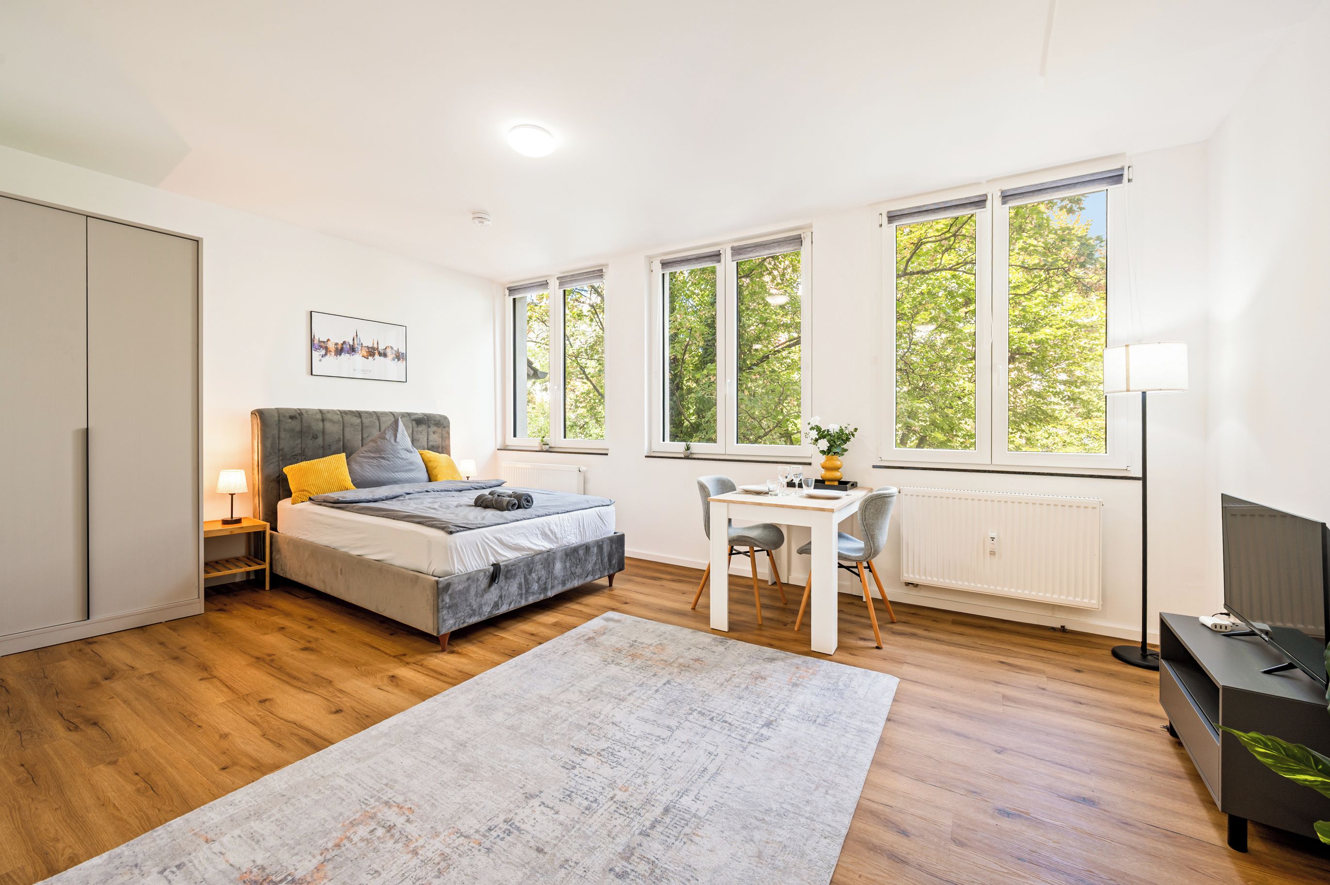 A modern bedroom with a bed, bedside table, lamp, and a desk with a chair. There are large windows providing natural light and a view of trees.