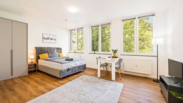 A modern bedroom with a bed, bedside table, lamp, and a desk with a chair. There are large windows providing natural light and a view of trees.