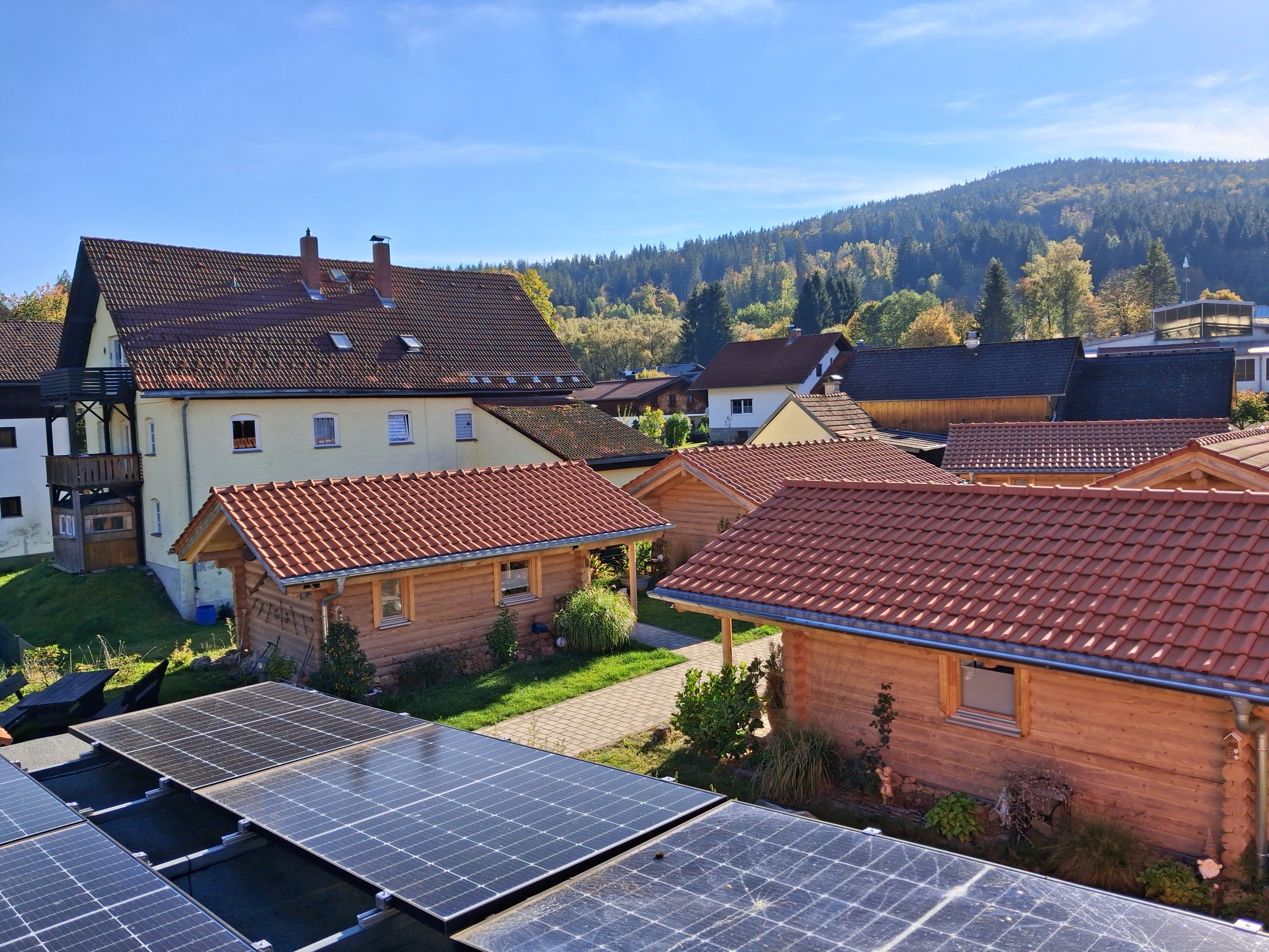 Ein Wohngebiet mit mehreren Häusern, von denen einige mit Solaranlagen auf den Dächern ausgestattet sind, und einem bewaldeten Hügel im Hintergrund.