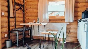 A wooden cabin interior with a dining table, chairs, a hanging light fixture, a radiator, and a window with curtains.