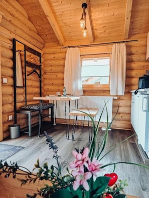 A wooden cabin interior with a dining table, chairs, a hanging light fixture, a radiator, and a window with curtains.