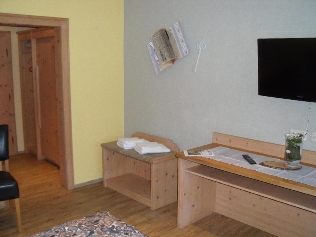 A small room with a wooden desk, a chair, a bed with a towel, a television mounted on the wall, and a small table with a remote control.