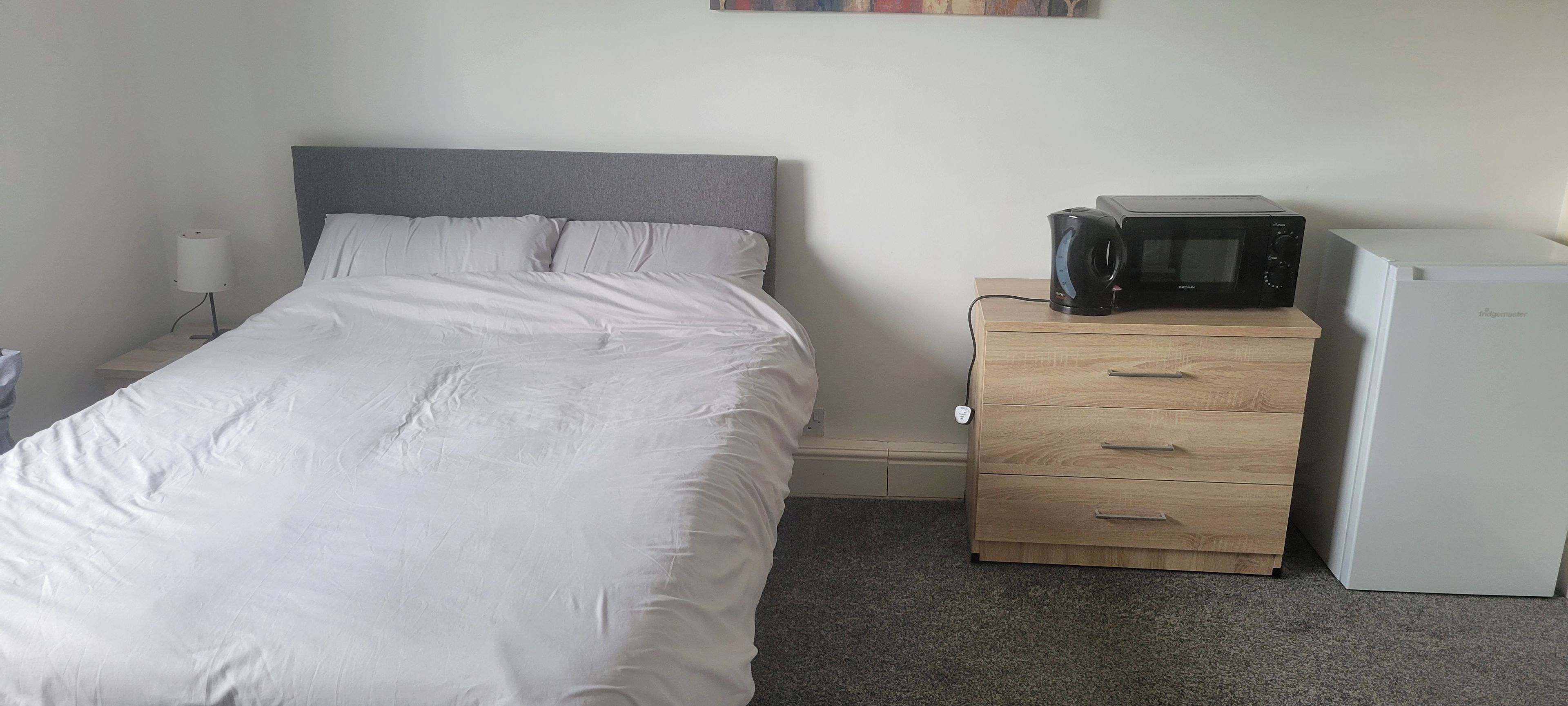 A bedroom with a bed, a bedside table, a microwave, and a refrigerator.