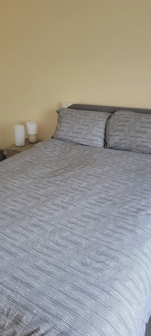 A bed with a striped bedspread and two pillows, a bedside table with a lamp, and a wall with a textured finish. - Apartment 27 Sleeps 2 With Enclosed Garden (Saltash)
