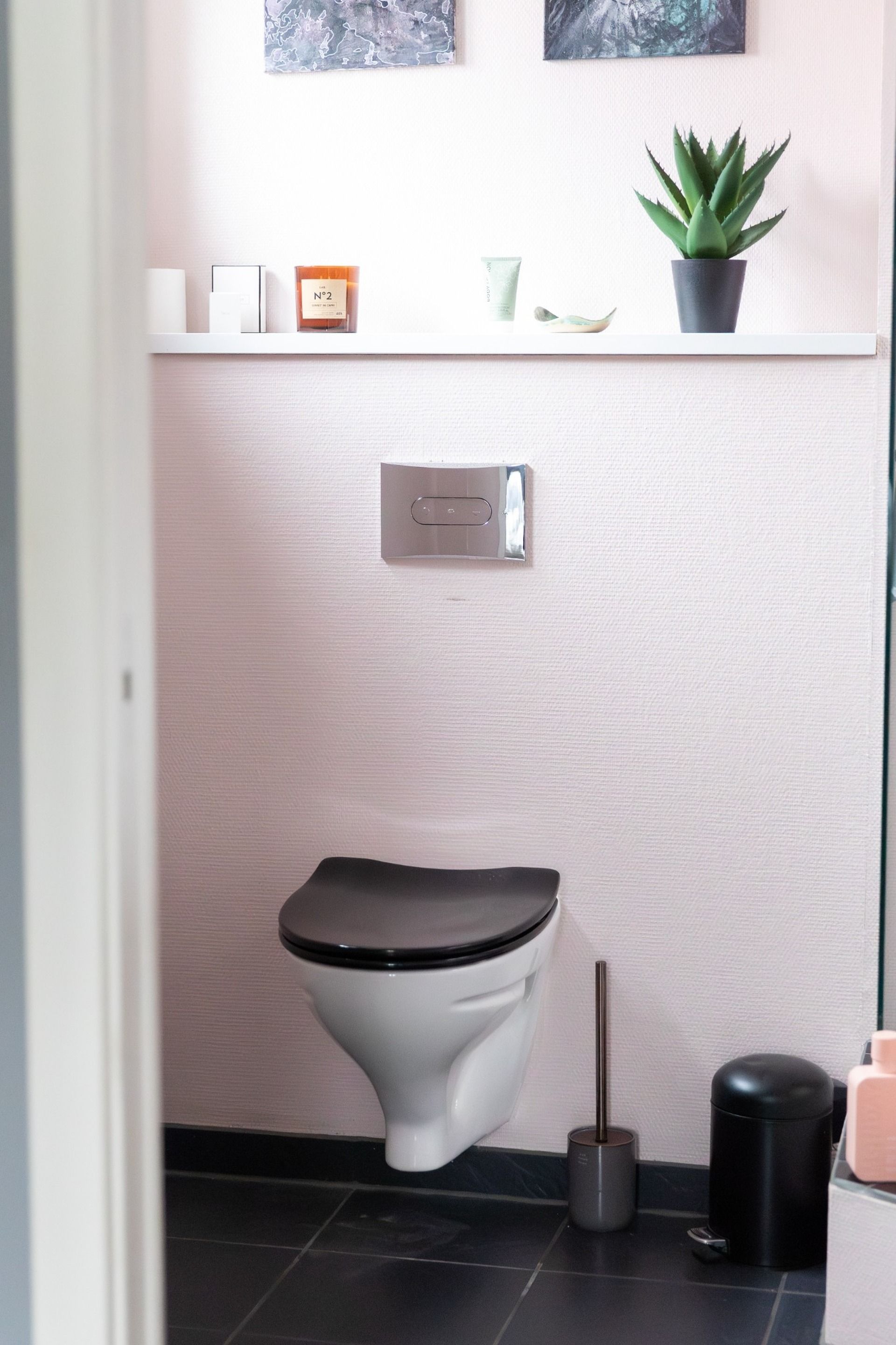 A modern bathroom with a toilet, a trash bin, a toilet brush, and a potted plant on a shelf.