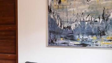 Un sofá gris con un cojín color melocotón y un objeto azul y blanco sobre una mesa blanca. Un cuadro abstracto de gran tamaño adorna la pared.