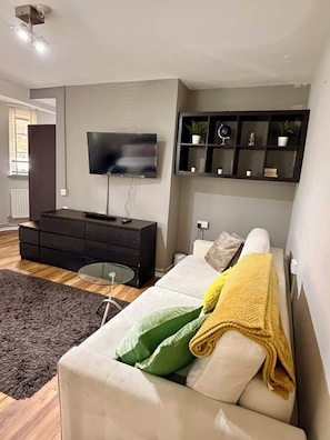 A modern living room with a flat-screen TV mounted on the wall, a dark wooden cabinet, a white sofa with green and yellow pillows, and a small glass table. - Cosy London Stay near Shoreditch (London)