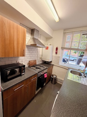 A kitchen with wooden cabinets, a built-in microwave, a stove, and a window with blinds. - Cosy London Stay near Shoreditch (London)