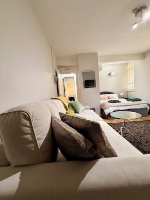 A modern living room with a sofa, a glass coffee table, and a bed in the background. - Cosy London Stay near Shoreditch (London)