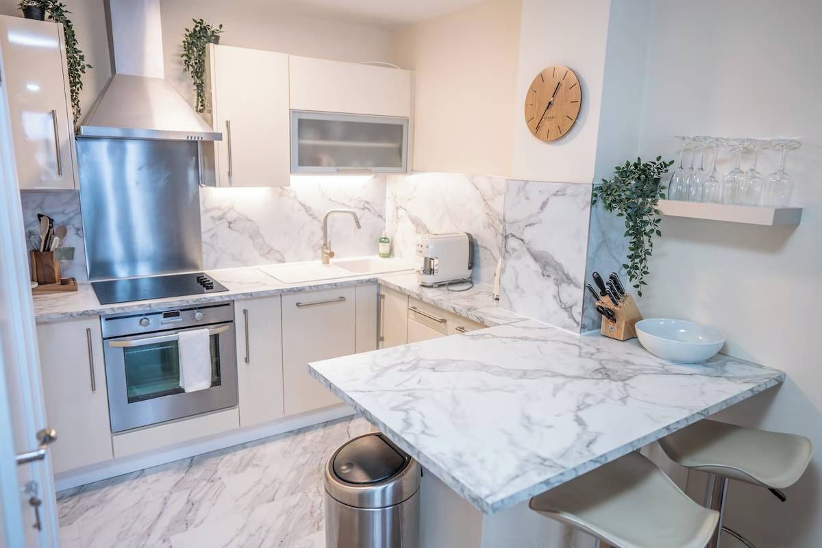A modern kitchen with white cabinetry, a stainless steel range hood, a built-in oven, a marble countertop, and a wall-mounted clock.