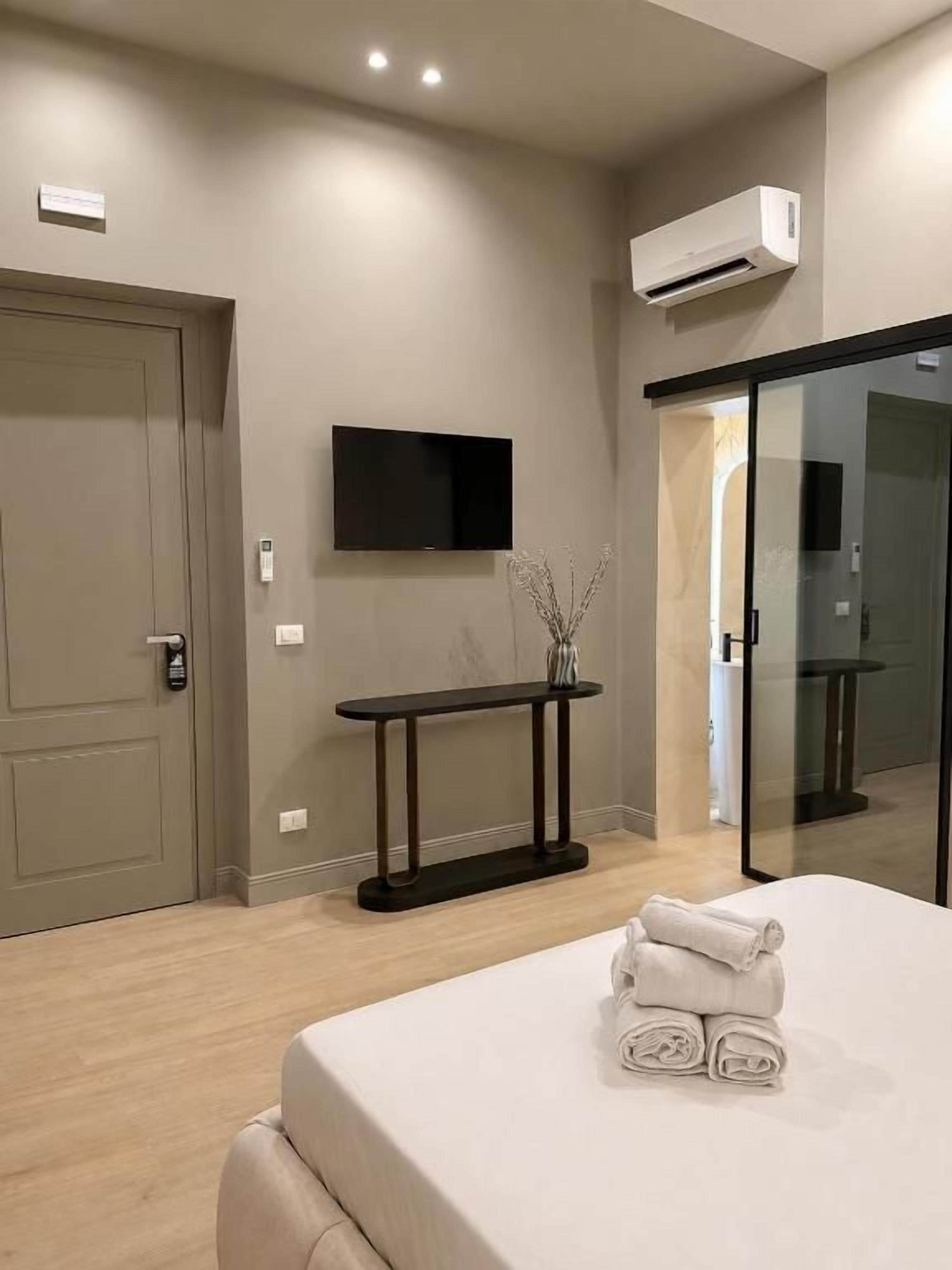 A hotel room with a bed, a flat-screen TV mounted on the wall, a bedside table, and a glass door leading to a bathroom.