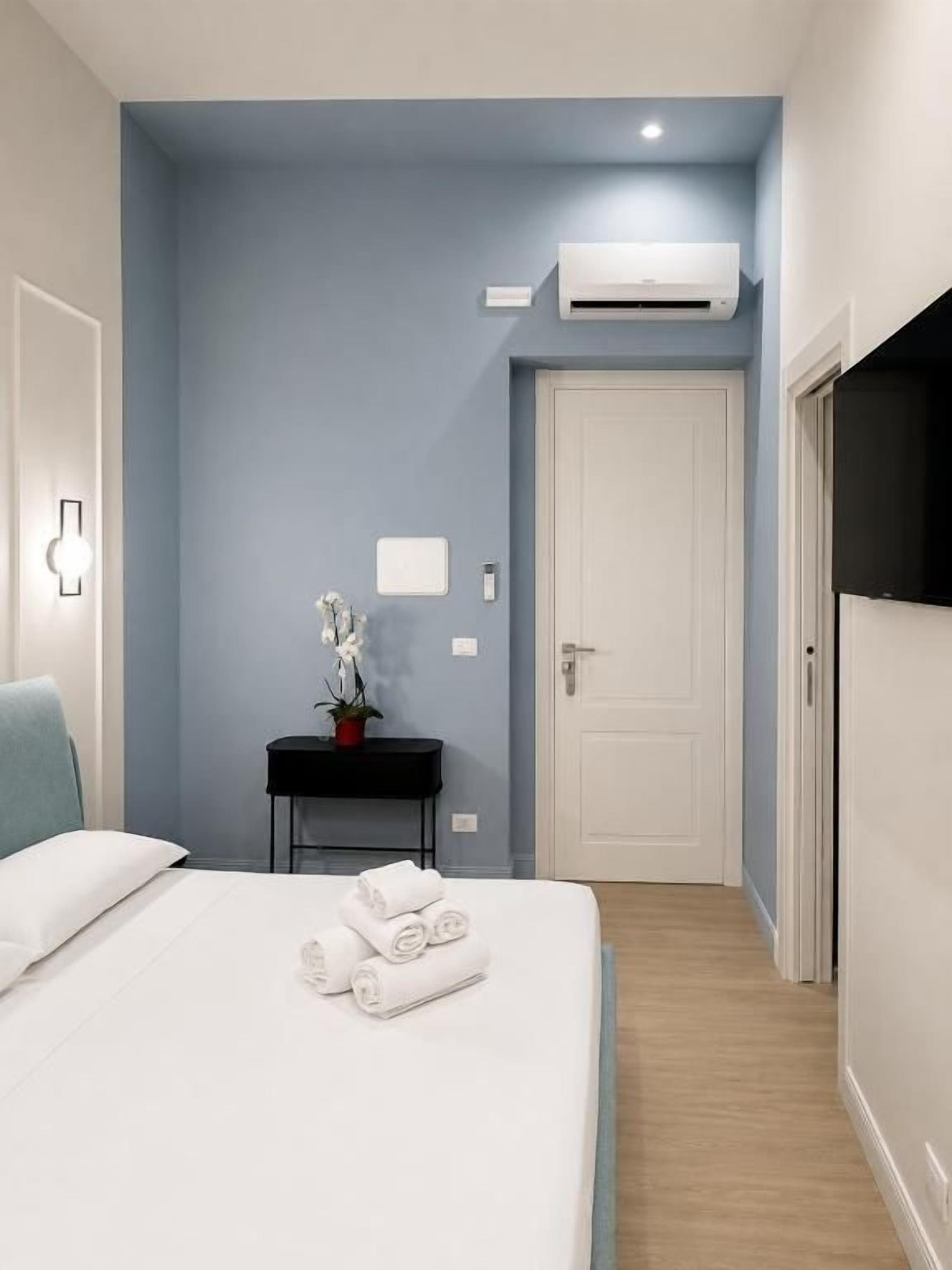 A hotel room with a bed, a bedside table with a lamp, a wall-mounted air conditioner, and a door.