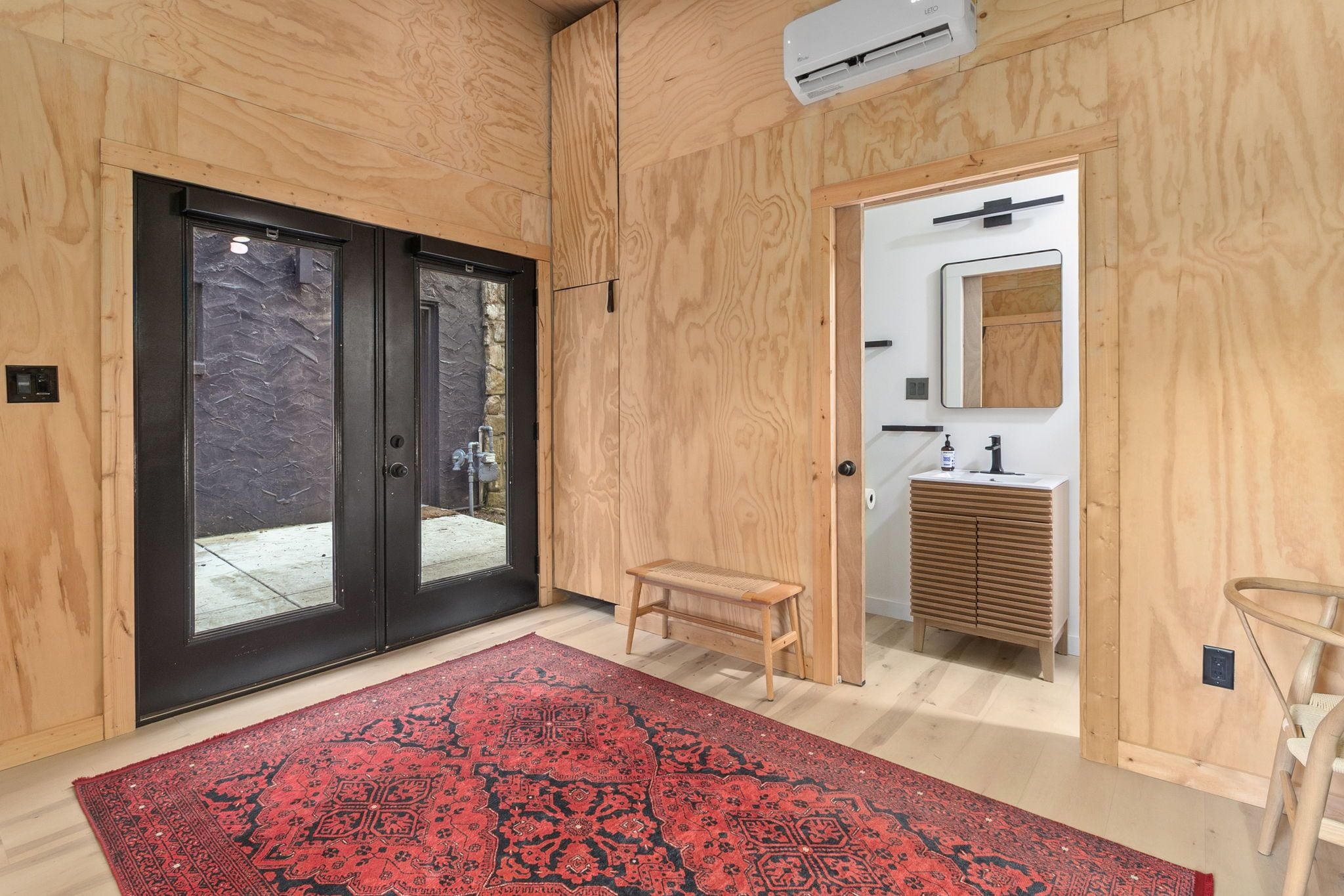 A modern interior with a wooden floor, a red patterned rug, a small wooden bench, and a bathroom with a sink and mirror.