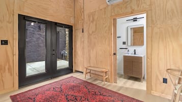 A modern interior with a wooden floor, a red patterned rug, a small wooden bench, and a bathroom with a sink and mirror.