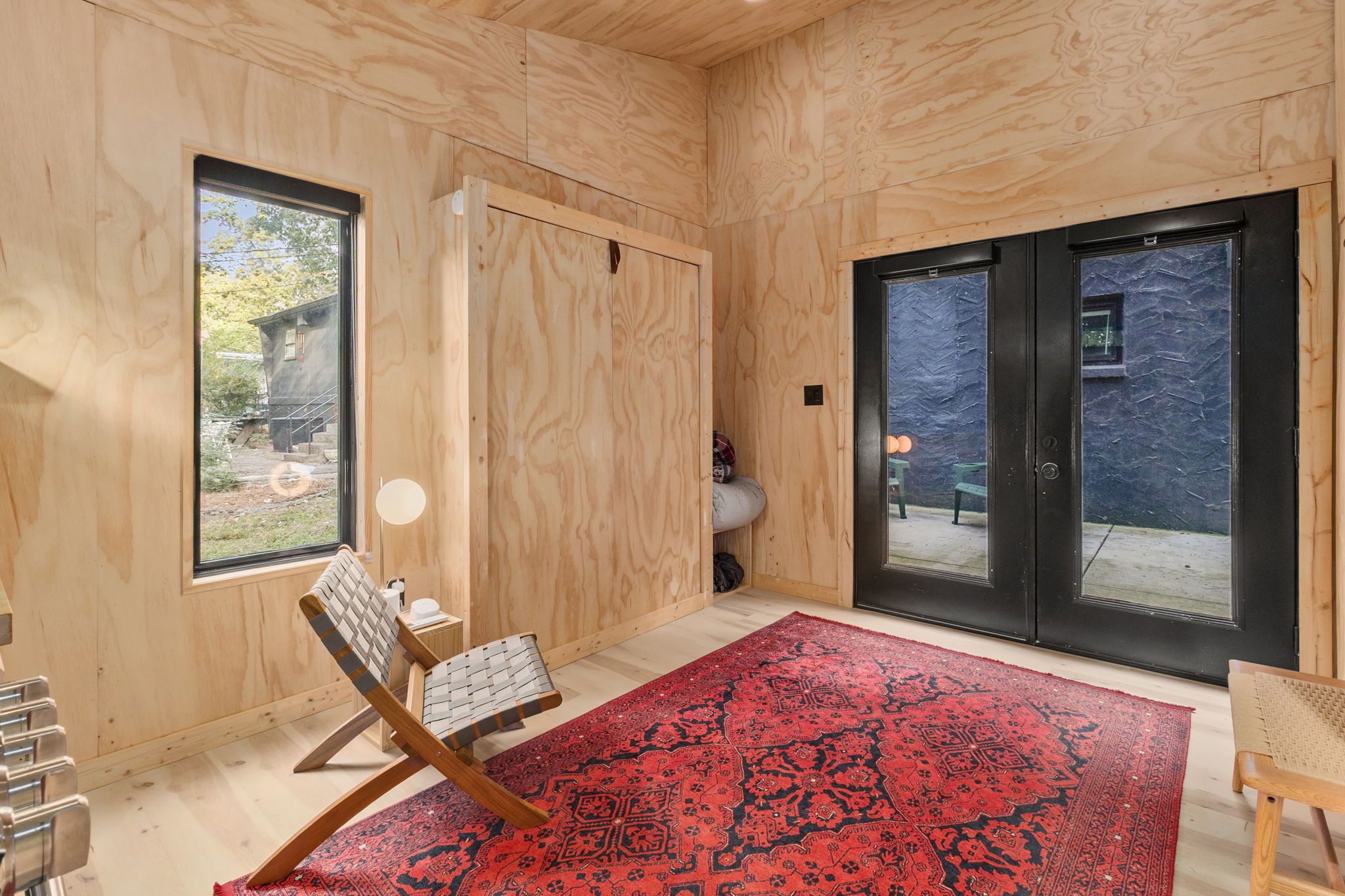 A room with wooden walls, a large window, a sliding door, a red patterned rug, and a chair.