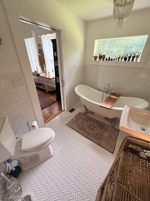 Ein Badezimmer mit einer freistehenden Badewanne, einer Toilette und einem Waschbecken. Ein Fenster bietet Blick auf Grünflächen draußen.