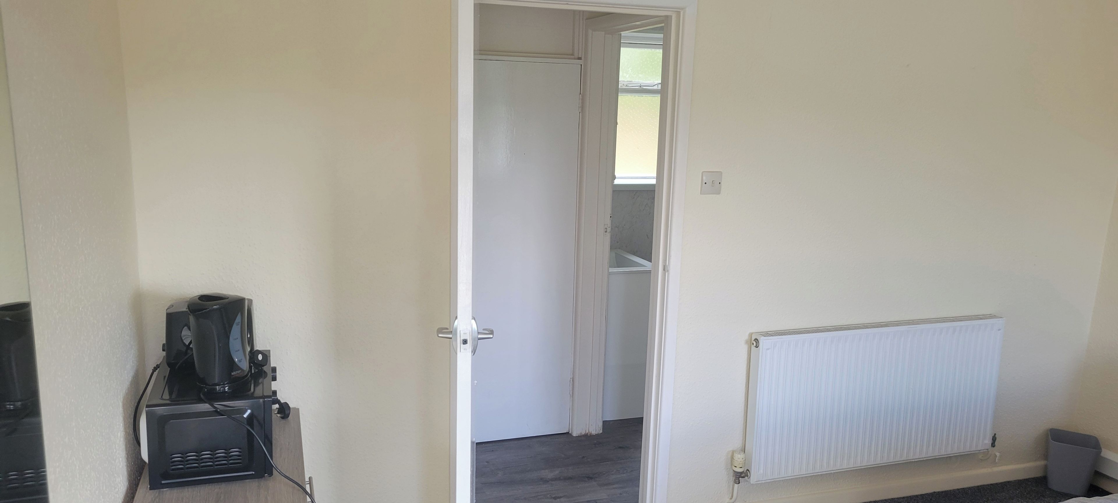 Una habitación con un radiador blanco, una puerta cerrada y una cafetera sobre un pedestal.