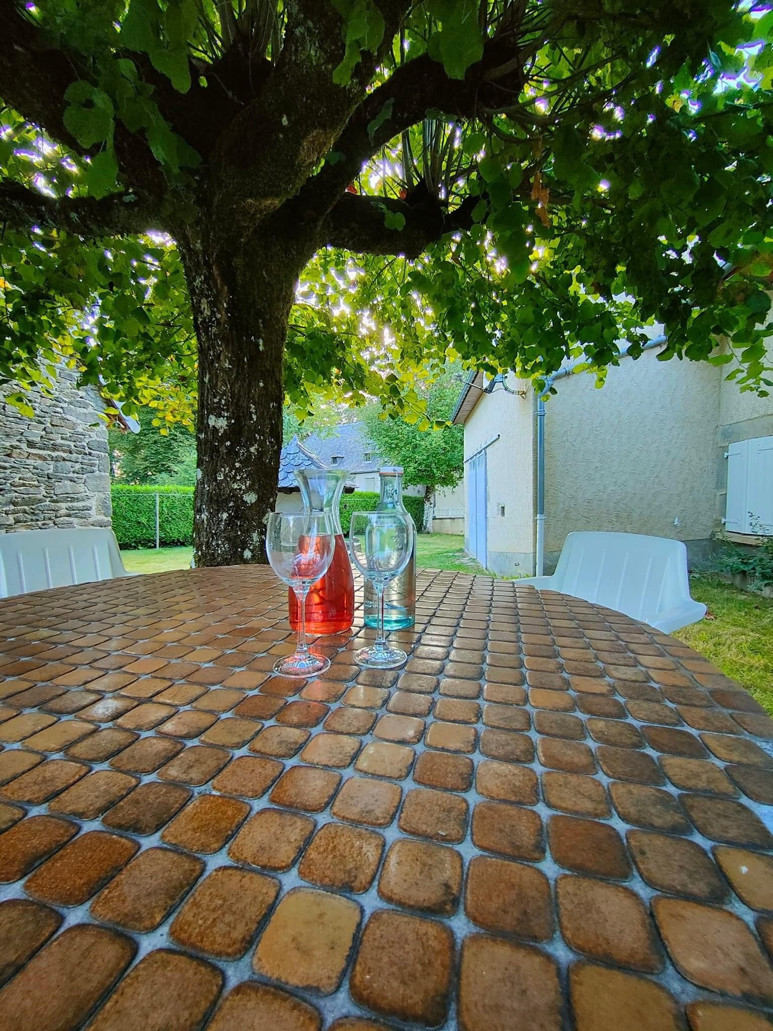 A brick table with four wine glasses and a red bottle, surrounded by a tree and a building.