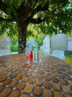 Een bakstenen tafel met vier wijnglazen en een rode fles, omringd door een boom en een gebouw.