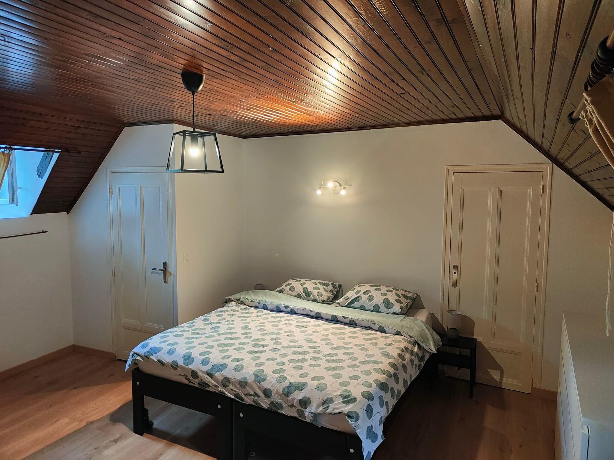 A bedroom with a sloped ceiling, a bed with patterned bedding, a bedside table, and a wall-mounted light fixture.